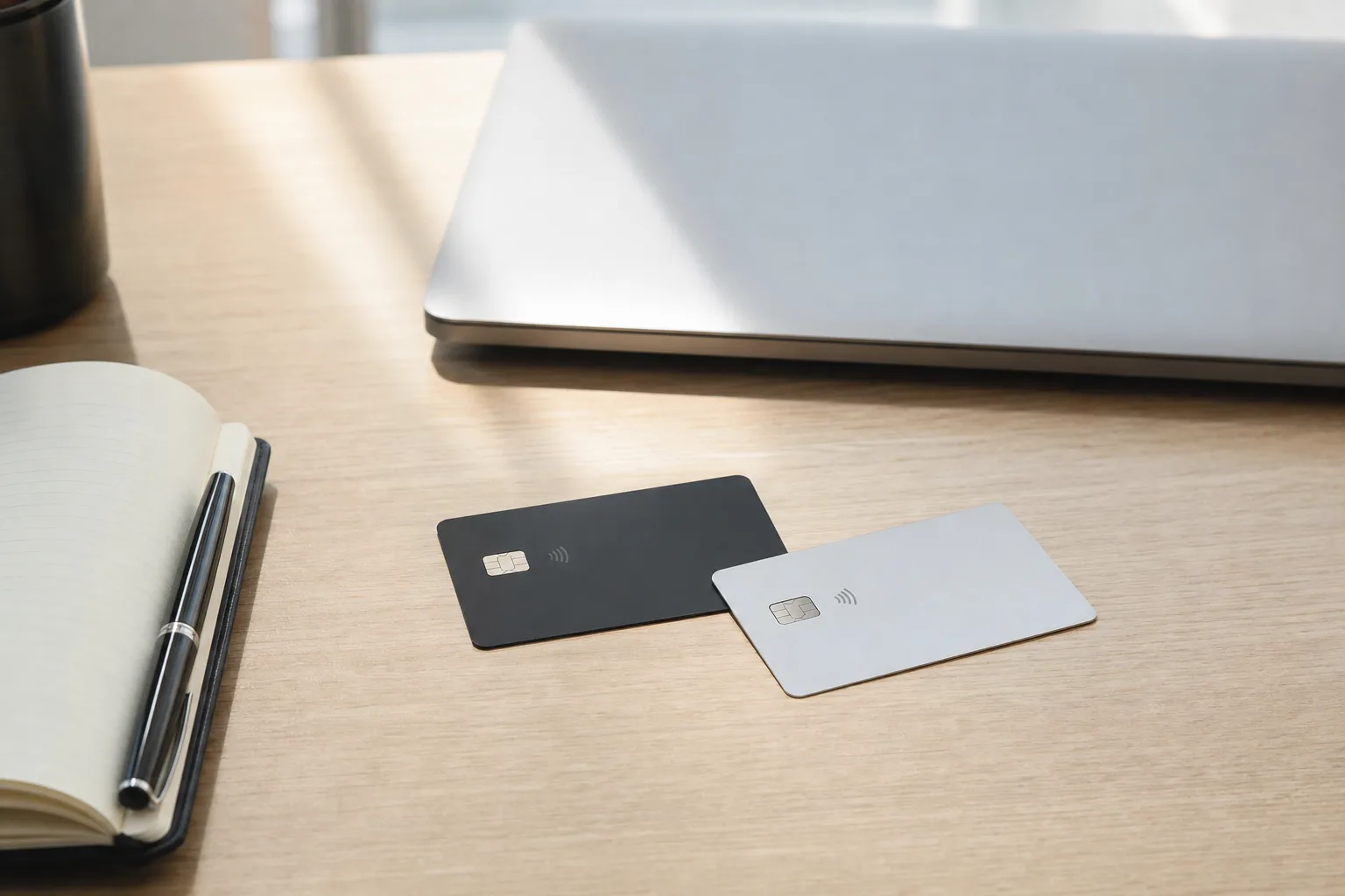 Deux cartes bancaires posées sur un bureau en bois clair à côté d'un carnet ouvert, d'un stylo et d'un ordinateur portable fermé